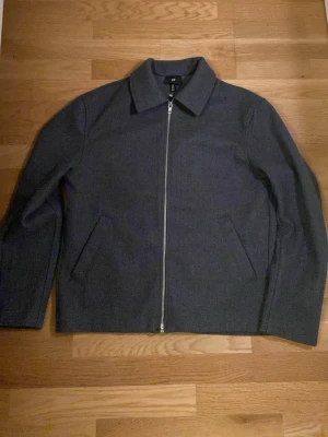 Grå ulljacka med dragkedja från H&M - Säljer en stilren grå jacka från H&M i ullblandning. Jackan har rak passform, klassisk krage och hel dragkedja framtill. Två snedställda fickor på sidorna och enkel design som funkar till många outfits.