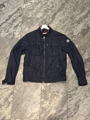 Svart vindjacka från Moncler - Säljer min mörkblåa jacka från Moncler med klassisk logga på ärmen. Jackan har dragkedja framtill, flera fickor med knappar och en hög krage med röd-vit detalj. Perfekt till vår, höst och en frisk sommarkväll. Använd sparsamt. Storlek 3 motsv. S/M hör av er vid frågor!
