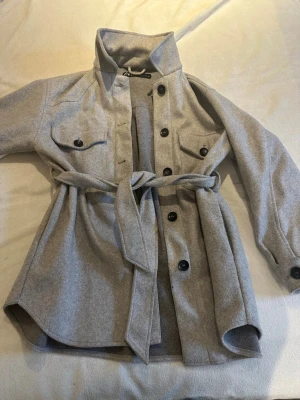 Grå vår jacka från Zara med bälte - Säljer en ljusgrå vår jacka från Zara med stora svarta knappar, två bröstfickor och ett matchande bälte i midjan. Jackan har klassisk krage och är långärmad, perfekt för lager på lager under höst och vår.