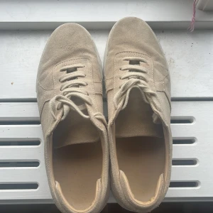 Beige sneakers från Axel Arigato mocka - Snygga beige sneakers från Axel Arigato i mjuk mocka med klassisk rund tå och vita snören. Skorna har en stilren design med diskret logga på sidan och en vit platt sula. Perfekta för dig som gillar minimalistisk och clean stil.