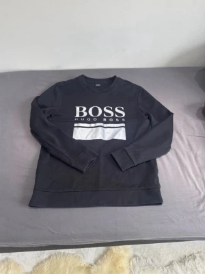 Svart sweatshirt från Hugo Boss L - Säljer en svart sweatshirt från Hugo Boss i storlek L. Tröjan har ett stort vitt BOSS-tryck på bröstet och en vit detalj under loggan. Klassisk rund halsringning och långa ärmar. Tillverkad i mjuk bomull för skön känsla.