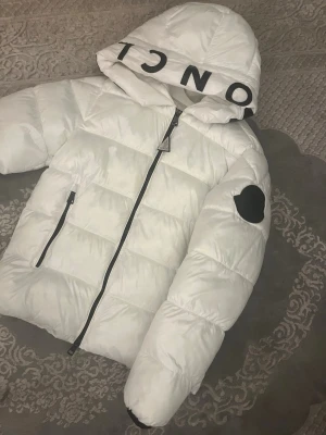 Vit Moncler pufferjacka med huva - Säljer en riktigt snygg vit pufferjacka från Moncler med stor huva och svart logga på ärmen. Jackan har dragkedja framtill, två sidofickor och stora svarta bokstäver på huvan. Perfekt för kalla dagar och ger en clean look. Den är i nyskicka knappt använd 