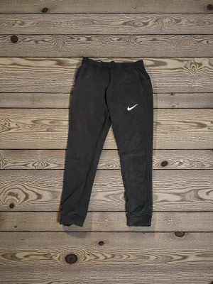 Svarta Nike Dri-FIT mjukisbyxor M - Svarta mjukisbyxor från Nike med Dri-FIT-teknologi och klassisk vit swoosh-logga på benet. Byxorna har resår i midjan, smal passform och muddar vid bensluten. Perfekta för chill eller träning.