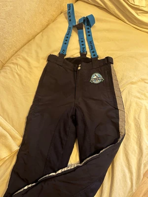 Ridtäckbyxor från Horze med blå detaljer - Täckbyxor från Horze i marinblått med blå detaljer och broderad patch 'Rider of the World'. Justerbara blå hängslen och reflexdetaljer på sidan. Kardborre vid benslut och förstärkta partier. Perfekta för kalla dagar och ridning. Använda några få gånger, som nya! 