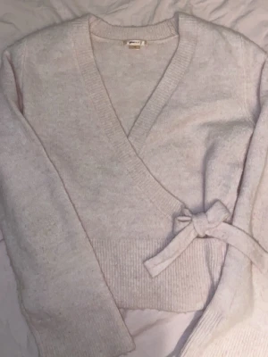 Ljusrosa omlottkofta från Gina Tricot - Supersöt ljusrosa omlottkofta från Gina Tricot med knyt i sidan. Koftan är stickad och har långa ärmar samt bred ribbad kant vid ärmslut och nederkant. Perfekt för lager på lager under höst och vinter!