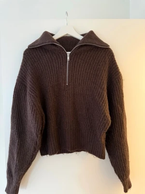 Brun stickad half zip från Gina Tricot - Mysig brun stickad tröja från Gina Tricot med bred krage och half zip-dragkedja framtill. Tröjan har en loose passform och långa ärmar, perfekt för kyliga dagar. Ribbstickad struktur ger en trendig och avslappnad look.