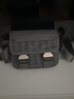 Lacoste Axelremsväska - Snygg Lacoste-väska i mörkgrått med geometriskt mönster och den välkända krokodillogotypen. • Skick: Nytt med etikett • Stängning: Flik med magnetisk tryckknapp och extra metallspännen •  Material: Högkvalitativt konstläder • Justerbar axelrem • Praktiska innerfack för telefon, plånbok och dokument  Perfekt för vardagsbruk - för arbete, skola eller resor.  Elegant, minimalistisk design i Lacostes sportiga stil