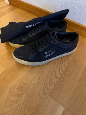 Svarta Saint Laurent sneakers - Säljer ett par svarta Saint Laurent sneakers med vit sula och handskriven logga på sidan. Skorna har snörning, rund tå och är tillverkade i en mix av canvas och skinn. Klassisk och clean design som passar till det mesta. Bra skick inga defekter. Det ända saken som man märker på är att en av slitningen (de ska vara med i designen) har blivit större än dem andra. Men det är inget hål eller nåt. Storlek 41. Bara dustbagen ingår. De är äkta.