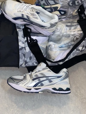 Vita och silver Asics sneakers - Säljer ett par fräscha Asics sneakers i vitt och silver med mörkblå detaljer. Skorna har meshpaneler för extra andningsförmåga och klassisk snörning. Perfekta för dig som gillar sportig stil och vill ha bekväma skor till vardagen.