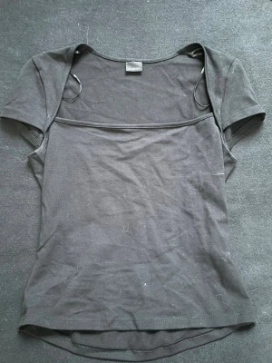 Svart kortärmad topp från Gina Tricot - En enkel svart topp från Gina Tricot med korta ärmar och rund halsringning. Tillverkad i mjuk bomullsmix och har en figurnära passform. Perfekt att matcha med jeans eller kjol för en clean look.