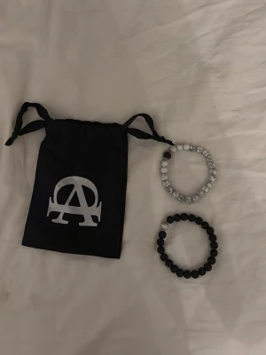 Två pärlarmband svart och vitmarmorerad - Säljer två stilrena pärlarmband, ett i mattsvart och ett i vitmarmorerad design. Båda har elastiskt band och detaljer i metall. Kommer med en svart tygpåse med stor A-logga. Perfekt accessoar för att lyfta din outfit.