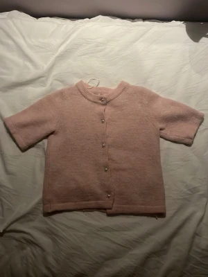 Ljusrosa kofta med kort ärm - Supersöt ljusrosa kofta med korta ärmar och rund hals. Koftan har pärlemorknappar framtill och är gjord i ett mjukt, stickat material som känns riktigt mysigt. Perfekt att slänga över en topp för en chill och söt look.