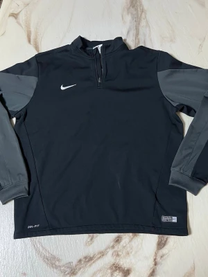 Nike Dri-Fit svart träningströja XL - Nike Dri-Fit träningströja i svart med mörkgrå ärmar och detaljer. Halv dragkedja framtill och broderad vit Nike-logga på bröstet. Tillverkad i funktionsmaterial som andas, perfekt för fotboll eller annan träning.