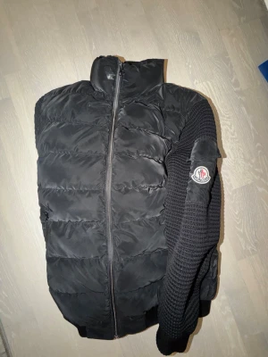 Svart pufferjacka från Moncler storlek S/M - Säljer en svart pufferjacka från Moncler med stickade ärmar och klassisk logga på ena armen. Jackan har dragkedja framtill och hög krage. Perfekt för dig som vill ha en stilren och varm jacka med snygga detaljer.
