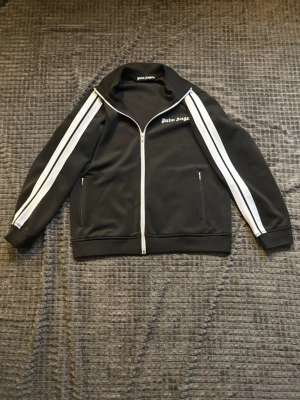 Palm Angels tröja svart - Säljer en svart tröja från Palm Angels med vita ränder längs ärmarna och logga på bröstet. Jackan har dragkedja framtill, hög krage och två fickor med dragkedja. Tillverkad i polyester för en sportig vibe.