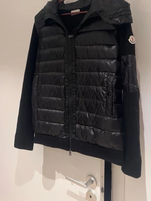 Svart dunväst från Moncler - Säljer en svart dunväst från Moncler med quiltad front och huva. Västen har dragkedja framtill, två fickor med knapp och klassisk Moncler-logga på ärmen. Materialmix med glansig polyester och stickade partier ger en sportig och clean look.