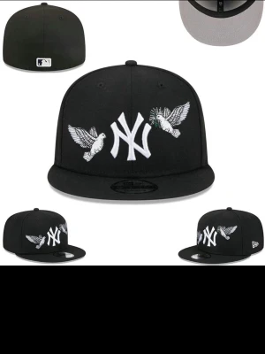 Svart New Era Yankees keps - Säljer en svart New Era keps med New York Yankees-logga framtill och två broderade vita duvor på sidorna. Klassisk flat peak och fitted passform. Perfekt för dig som vill ha en stilren och sportig look. Finns vilken size du vill