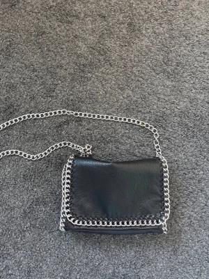 Svart axelväska med kedjedetaljer Tiamo - Säljer en svart axelväska från Tiamo i skinnimitation med silverfärgade kedjor runt kanterna och som axelrem. Väskan har dragkedja på baksidan och magnetknäppning framtill. Perfekt storlek för det viktigaste och ger en cool vibe till din outfit.