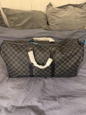 Svart Louis Vuitton keepall väska - Säljer en stor och rymlig resväska från Louis Vuitton i klassiskt svart och grått rutigt mönster. Väskan har dubbla handtag, silverfärgade metalldetaljer och dragkedja. Materialet är slitstark syntet med detaljer i skinn.