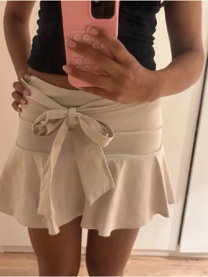 Beige kortkjol från Zara med volang och rosett - Supersöt beige kortkjol från Zara i storlek Small. Kjolen har bred midja och stor knytrosett framtill. Kjolen har volangkant som ger en extra feminin touch och är tillverkad i ett mjukt tyg som känns bekvämt. Perfekt för dig som gillar en söt och trendig stil.💞💞