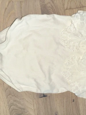 Vit väst med spetsdetaljer från Zara Zara vit body, storlek 80 - Vit body med spetsdetaljer från Zara. Säljer en söt vit väst från Zara i ribbad bomull med dekorativa spetsapplikationer på framsidan. Perfekt för att addera en romantisk touch till outfiten. Passar barn i storlek 80, 9-12 månader.