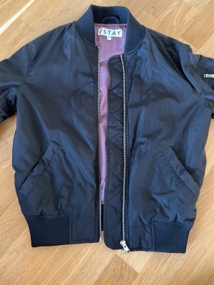 Svart bomberjacka från Stay XS - Snygg svart bomberjacka från Stay i storlek XS. Jackan har en klassisk bombermodell med ribbade muddar, silverfärgad dragkedja och fickor på sidorna. Insidan är fodrad med ett ljusrosa tyg som ger en cool kontrast. Perfekt till vardagslooken.