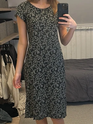 Svart blommig klänning från KappAhl - Supersöt svart klänning från KappAhl med små vita blommor över hela tyget. Modellen är knälång med korta ärmar och rund halsringning. Perfekt för dig som gillar en enkel men ändå söt stil. Passar också som nattlinne och är väldigt skön att ha på sig :)