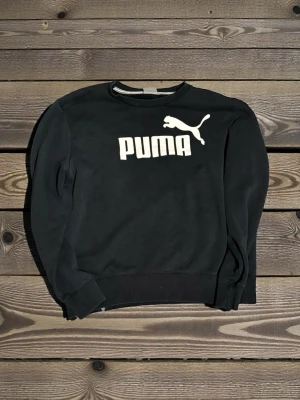 Svart sweatshirt från Puma - Svart sweatshirt från Puma i storlek S med klassisk vit logga och text framtill. Tröjan har rund halsringning, ribbade muddar vid ärmslut och nederkant samt lång ärm. Perfekt för en chill och sportig look.