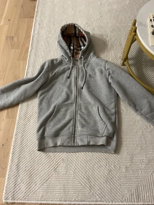 Grå hoodie från Burberry med dragkedja - Snygg grå hoodie från Burberry med klassiskt rutigt foder i huvan. Tröjan har dragkedja framtill, två fickor och justerbar huva med snören. Perfekt för en stilren och avslappnad look. Materialet är mjukt och bekvämt, perfekt för vardagsbruk.