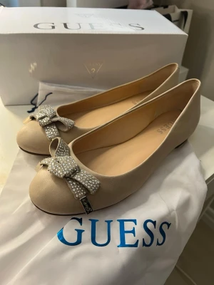 Beige ballerinaskor med rosett Guess - Snygga beige ballerinaskor från Guess med en stor glittrig rosett framtill, dekorerad med små stenar och metallplatta med logga. Skorna har rund tå och är tillverkade i ett mjukt material som ger en elegant look. Perfekta för dig som vill ha något extra till outfiten.