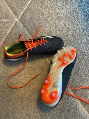  Adidas predators - Säljer ett par snygga fotbollsskor från Adidas i svart med vita ränder och orange detaljer. Skorna har orange snörning, grön insida och genomskinlig sula med orange dobbar. Perfekta för dig som vill sticka ut på planen! Storlek 40. Är i fint skick fast ändå använda.