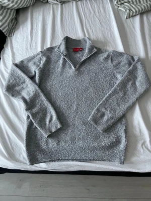 Grå stickad halvzip-tröja - Mysig grå stickad tröja med halv dragkedja och hög krage. Tröjan har lång ärm och ribbade muddar vid ärmslut och nederkant. Perfekt för lager-på-lager och chill dagar. Materialet ser ut att vara mjukt och bekvämt.