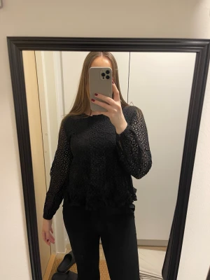 Svart spetsblus med lång ärm - Säljer en snygg svart blus i spets med långärmade ärmar och rund halsringning. Blusen har ett diskret blommönster i spetsen och volangdetaljer nedtill. 