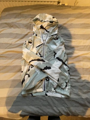 Vit camo vindjacka med huva - Snygg vindjacka i vitt med svart och grått camomönster. Jackan har dragkedja framtill och två fickor med dragkedja.  Pris ej hugget i sten 