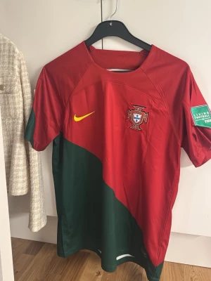 Portugal Ronaldo 7 Nike fotbollströja - Säljer en Portugal landslagströja från Nike med Ronaldo 7 på ryggen. Tröjan är röd med gröna detaljer och gula tryck, har FIFA World Cup Qatar 2022 och Living Football FIFA patchar på ärmarna. Tillverkad i polyester med Dri-FIT teknologi för extra komfort.