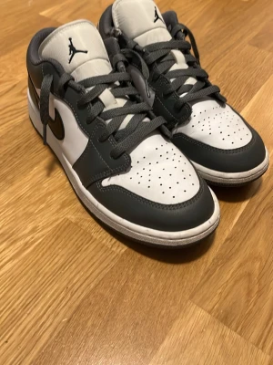 Air Jordan 1 Low grå/vit - Snygga Air Jordan 1 Low sneakers i grå och vitt läder med klassisk Jumpman-logga på plösen och svart swoosh på sidan. Skorna har perforerad tåbox för ventilation och gråa skosnören. Perfekt för dig som gillar streetwear och ikonisk design. Har några tecken på användning men annars så är de bra.