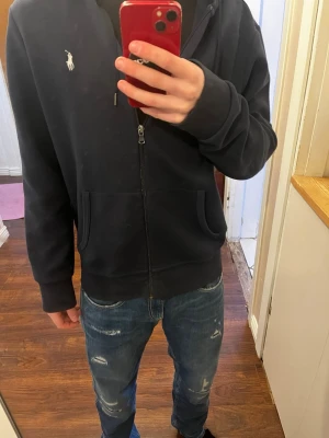 Mörkblå hoodie från Polo Ralph Lauren - Snygg mörkblå hoodie med dragkedja från Polo Ralph Lauren. Klassisk design med vit logga på bröstet. Tillverkad i mjukt bomullsmaterial som är skönt att bära. Storlek M 175 självklart äkta hör av er vid frågor 