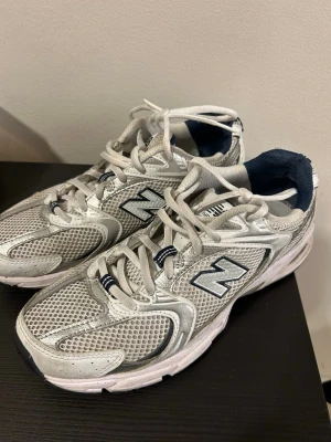 New Balance 530 vita sneakers - Snygga New Balance 530 sneakers i vitt och silver med marinblå detaljer. Ovandelen är i mesh och syntet för bra andningsförmåga och komfort. Klassisk retrostil med tydlig logga på sidan och bekväm sula med ABZORB-dämpning.