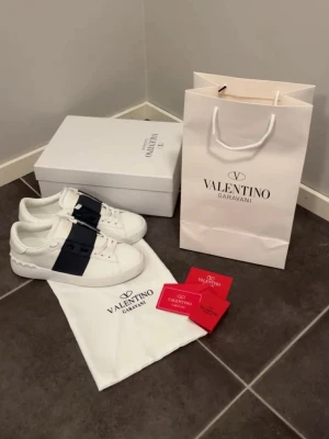 Valentino Garavani vita sneakers - Snygga vita sneakers från Valentino Garavani med bred marinblå rem över snörningen och diskreta nitar på hälen. Tillverkade i slätt läder med klassisk låg profil och rund tå. Perfekta för dig som vill ha en stilren och exklusiv look. Originalkartong och äkthetskort medföljer! Skorna köptes här på plick men säljs vidare pågrund av för liten storlek.                               Hör av dig vid frågor och funderingar!