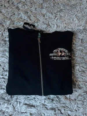 Svart hoodie från Ralph lauren - Svart hoodie med dragkedja från Sportsworld. Broderad logga och text på bröstet, två framfickor och justerbar huva med snören. Tillverkad i mjukt material, perfekt för chill dagar eller när du vill ha en avslappnad look.