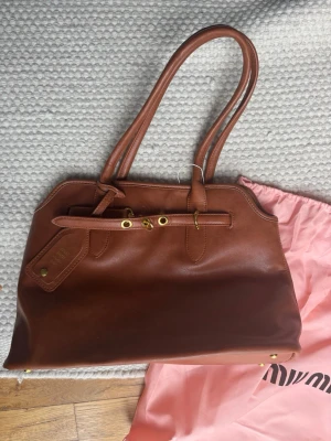 Brun väska från Miu Miu - Snygg brun väska från Miu Miu i mjukt läder med dubbla axelband och guldfärgade metalldetaljer. Väskan har en klassisk, rektangulär form och en tag med präglad logga. Perfekt för dig som gillar stilrena accessoarer med lyxig känsla. Du vet varför priset är lågt.