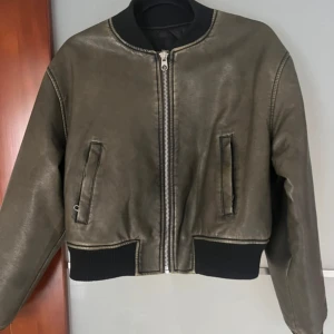 Olivgrön bomberjacka i fuskskinn - Snygg olivgrön bomberjacka i fuskskinn med svarta ribbade muddar vid ärmslut och midja. Jackan har dragkedja framtill, två snedställda fickor och en klassisk bomberkrage. Perfekt för dig som vill ha en stilren och trendig look.