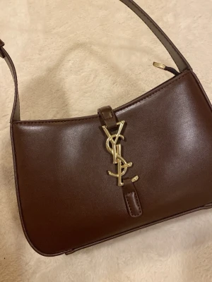 Brun axelremsväska från YSL i läder - Helt ny brun handväska från YSL i slätt läder med guldigt YSL-logo framtill. Väskan är köpt av en annan tjej och har därmed varken kvitto eller box. Kan inte vara säker på att den är äkta därav priset.