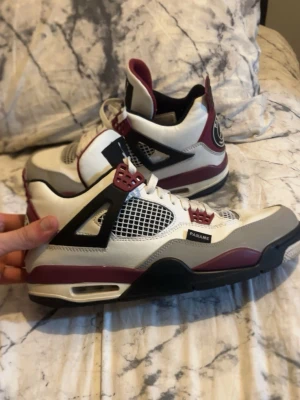 Air Jordan 4 PSG sneakers vit/bordeaux - Snygga Air Jordan 4 Paris Saint-Germain sneakers i vitt, bordeaux och svart. Skorna har meshpaneler, detaljer i läder och syntet, samt coola PSG-loggor på hälen. Klassisk snörning och ikonisk siluett som sticker ut.