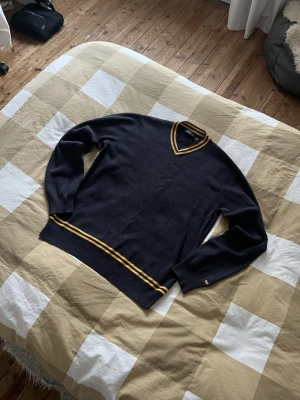 Ralph lauren knitwear  - Snygg mörkblå stickad tröja från Ralph Lauren med V-ringning och gula ränder vid halsringning och nederkant. Tröjan har långa ärmar och ribbstickade muddar. Perfekt för en sportig och stilren look. En liten skavank vid halsen men det går att sy annars är det ändå inget stort problem!