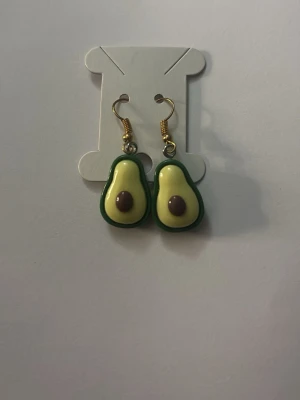 Avokado örhängen i plast - Säljer ett par coola örhängen formade som avokadohalvor. De är gjorda i plast med grön kant, ljusgul insida och brun kärna. Krokarna är guldfärgade och ger en lekfull touch till din stil. Perfekta för dig som gillar unika accessoarer!