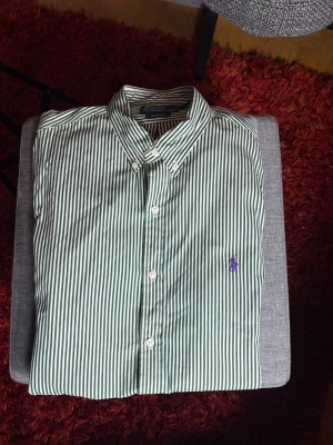 Randig grön skjorta från Polo Ralph Lauren - Snygg grön- och vitrandig skjorta från Polo Ralph Lauren i slim fit. Skjortan har klassisk krage, knappar framtill och den ikoniska lila loggan broderad på bröstet. Tillverkad i bomull och passar perfekt till en clean och stilren look.