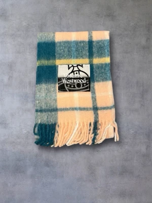 Vivienne Westwood halsduk  - Säljer en snygg rutig halsduk från Vivienne Westwood i mjuk ullblandning. Färgerna är beige, blågrön och inslag av gul, med klassiskt rutmönster och fransar i kanterna. Perfekt accessoar för att lyfta din outfit.