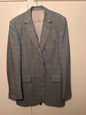 Grå rutig kavaj med klassisk passform från Tiger of Sweden - Stilren grå kavaj med diskret rutmönster och två knappar framtill. Klassisk passform med två fickor med lock och en bröstficka. Fodrad insida och smala slag ger en tidlös look. Perfekt för dig som vill ha en clean och dressad stil.