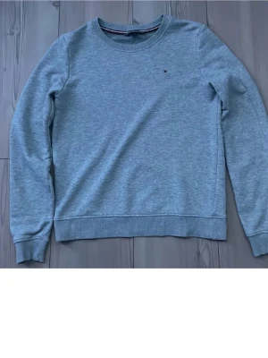 Ljusgrå sweatshirt från Tommy Hilfiger - Ljusgrå sweatshirt med rund hals från Tommy Hilfiger. Tröjan har en liten broderad logga på bröstet och ribbade muddar vid ärmslut och nederkant. Perfekt för en avslappnad och stilren look.